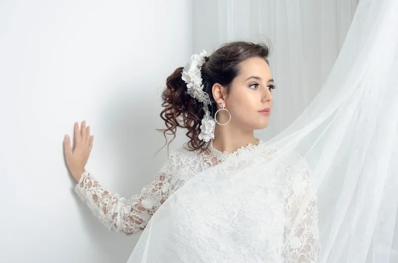 Close-up bridal portrait of model in white lace high-neck gown with floral hair accessories and flowing tulle veil