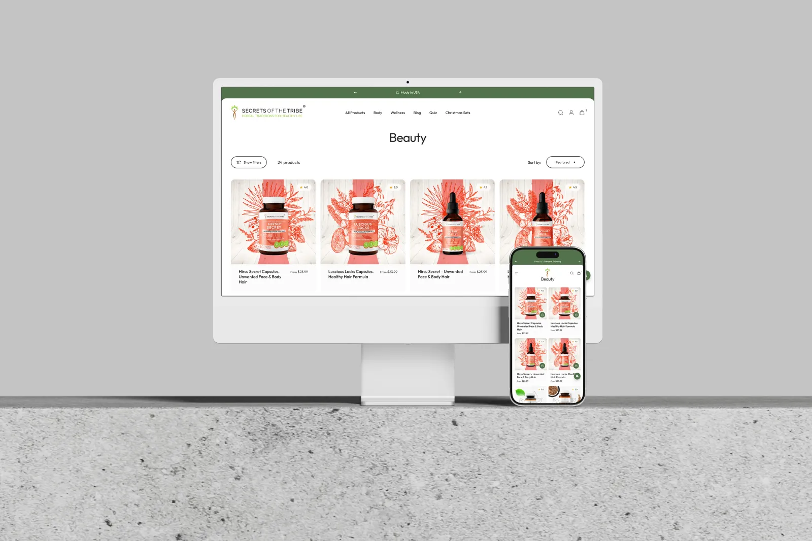 Desktop iMac monitor and iPhone side by side displaying the redesigned Secrets of the Tribe Shopify store Beauty collection page with herbal supplement tincture bottles in pink botanical packaging, set on a gray concrete surface